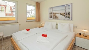 A neatly made double bed with white linens and red flowers, a bedside table with a lamp, and a window with a view of buildings. - Dune Residence C 13 - DR C 13 (Heringsdorf)
