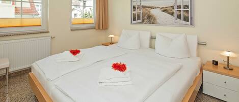 A neatly made double bed with white linens and red flowers, a bedside table with a lamp, and a window with a view of buildings.