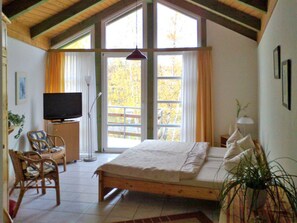 Un dormitorio con cama, televisor, una silla, planta y ventanas amplias.