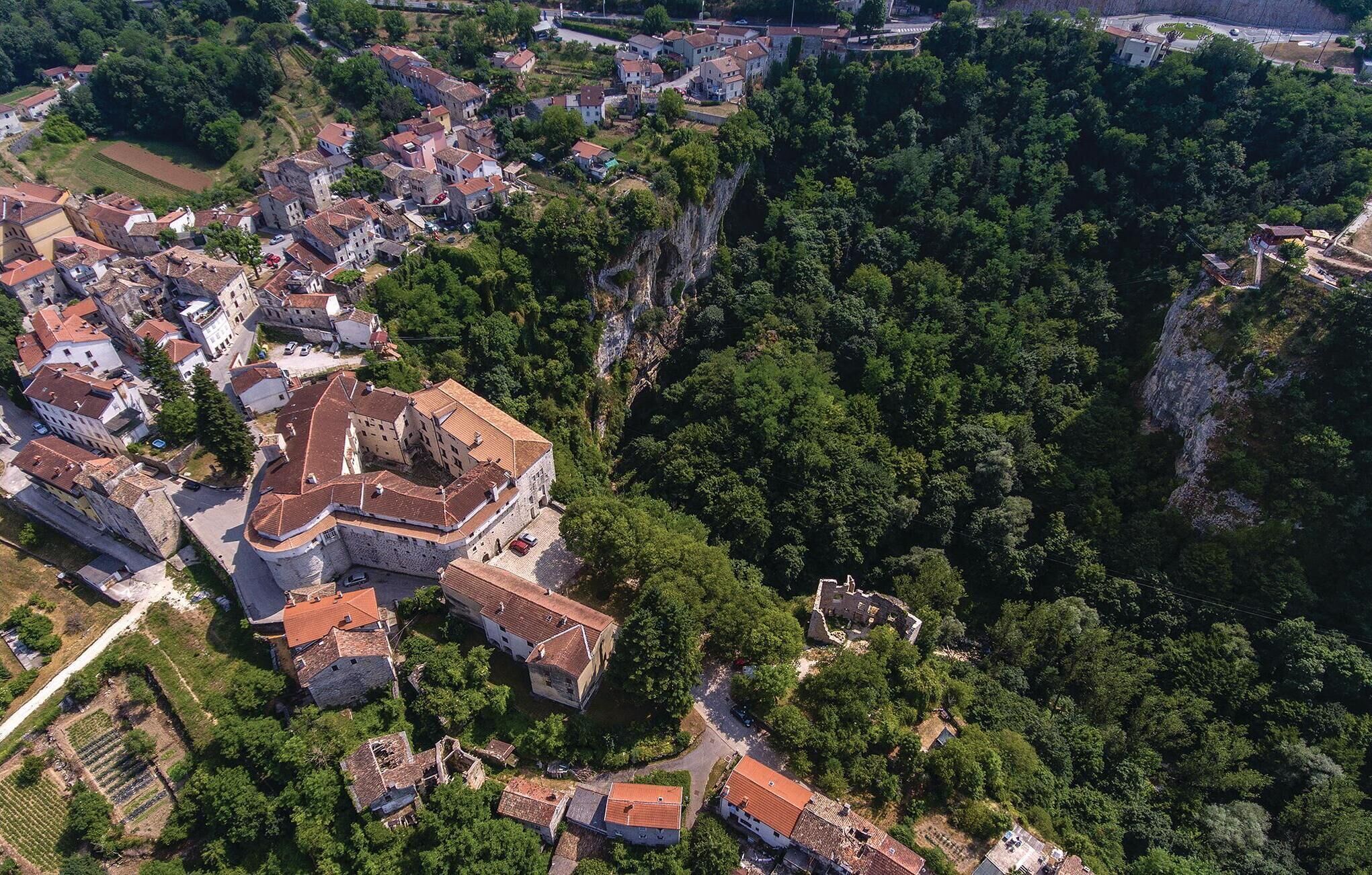 An aerial view of a historic town with a prominent fortified structure surrounded by residential buildings and lush greenery.