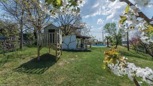Une cabane en bois avec une échelle, un petit bâtiment blanc et une piscine dans un jardin verdoyant.