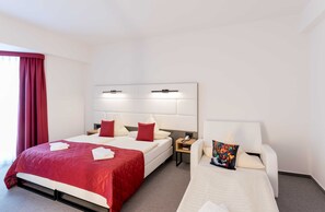 Une chambre d’hôtel avec deux lits, une grande fenêtre avec des rideaux rouges, une table de chevet avec une lampe et une petite table de nuit avec un téléphone.