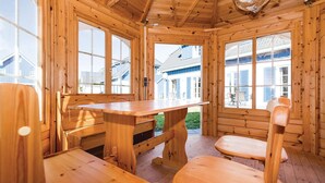 A wooden cabin with a large table and benches, large windows, and a view of a blue building outside. - Ferienhaus 5 Altefähr (Altefähr/Rügen)
