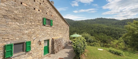 A stone building with a paved patio, green shutters, and a view of a forested hillside.