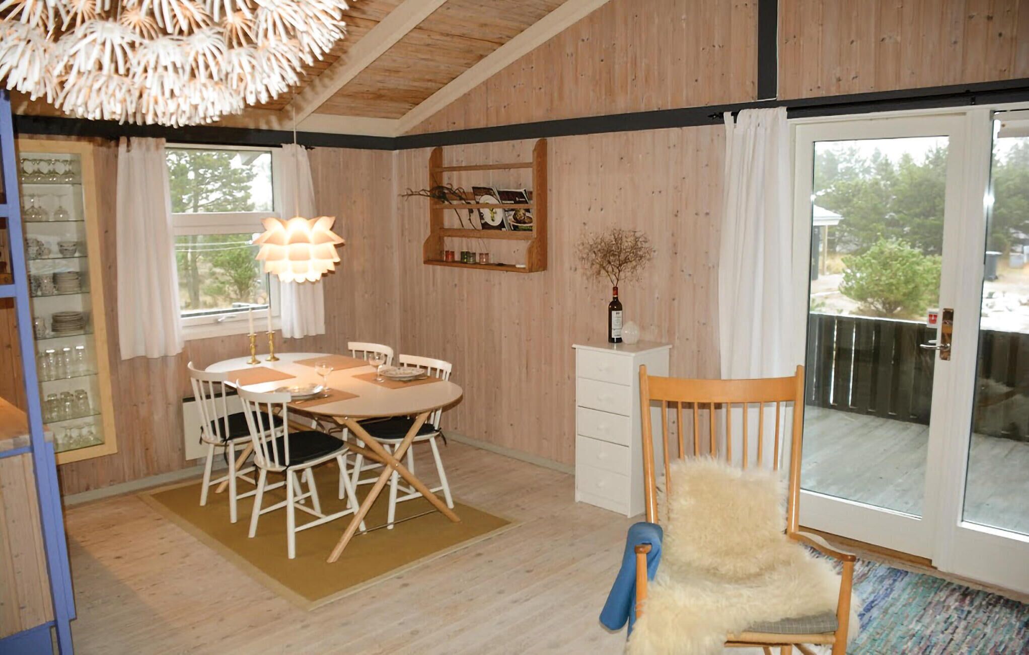 A dining area with a wooden table and chairs, a hanging light fixture, a white cabinet, and a view through a glass door.