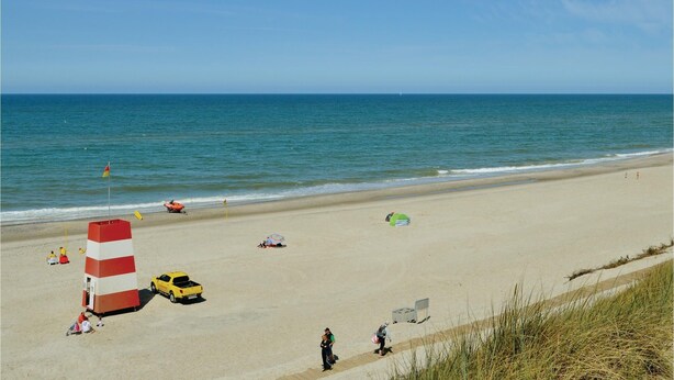 A beach with a lifeguard tower, a yellow vehicle, and people walking. - Cozy home in Ringkøbing with kitchen (Ringkøbing)