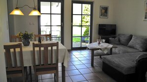 A living room with a sofa, a TV, a dining table with chairs, and a window with a view of greenery. - Holiday 2 - Holidays from the I Fam. Roman Kurth / TZR (Putbus)