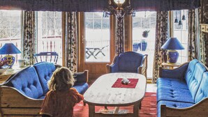 Una habitación acogedora con un ventanal grande, dos sillones azules, una mesa blanca y una alfombra roja.