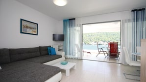 Una sala moderna con un sofá gris, una mesa de centro blanca y un televisor sobre un mueble blanco. Hay un balcón con vistas a un lago y colinas.
