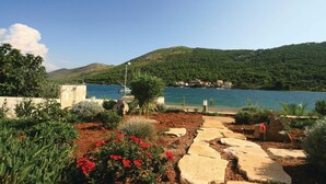 Un jardín costero con senderos de piedra, flores rojas y una vista de un lago con barcos y casas al fondo.