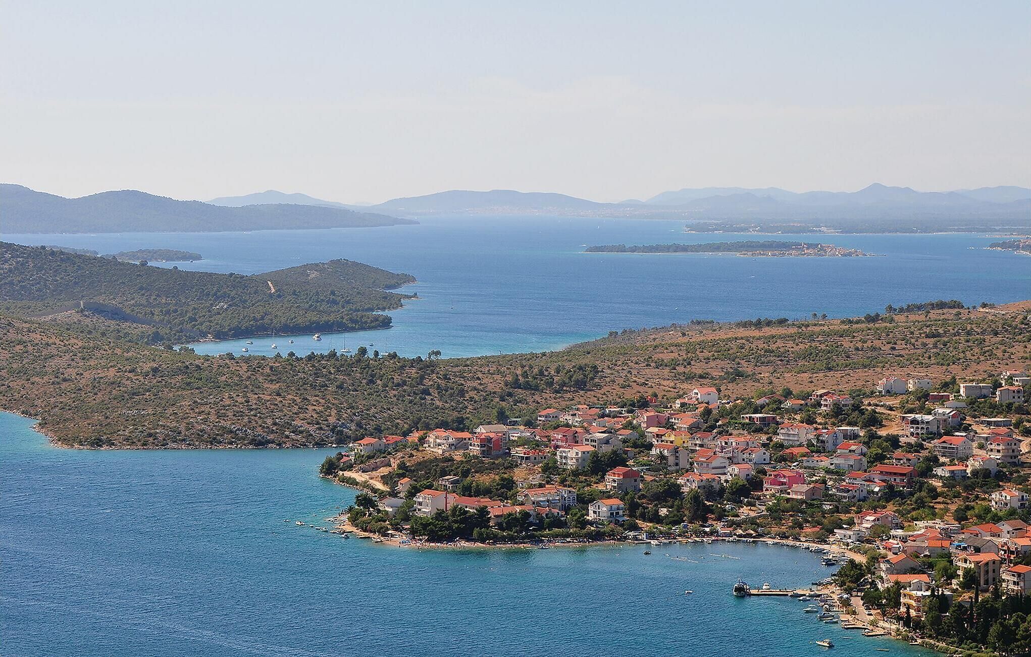 Une ville côtière avec des bâtiments qui bordent le littoral, une petite île avec des bateaux et, en arrière-plan, un paysage montagneux.