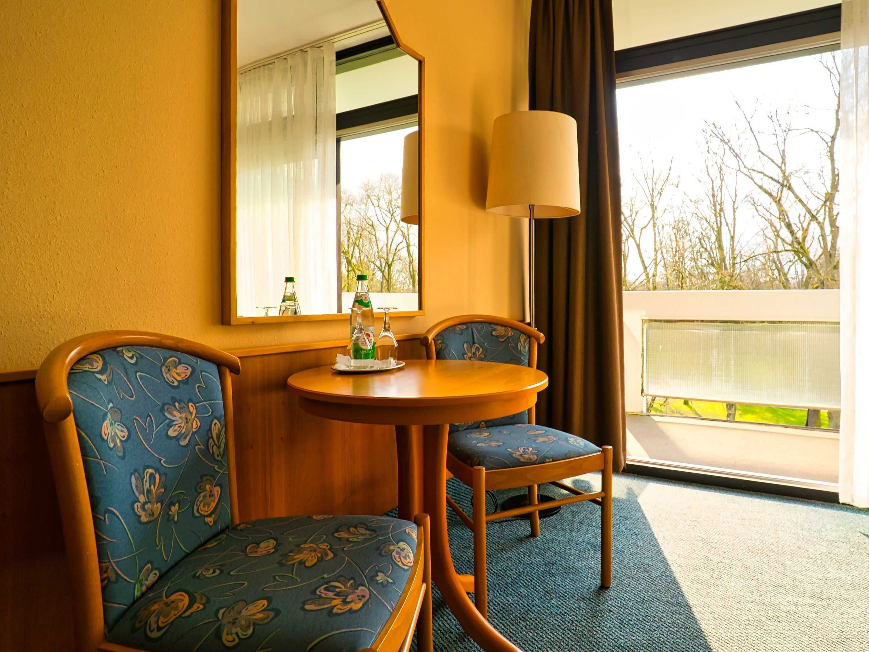 Une chambre d’hôtel avec une table en bois, deux chaises recouvertes d’un tissu à motifs floraux, un miroir et une vue sur les arbres par la fenêtre.