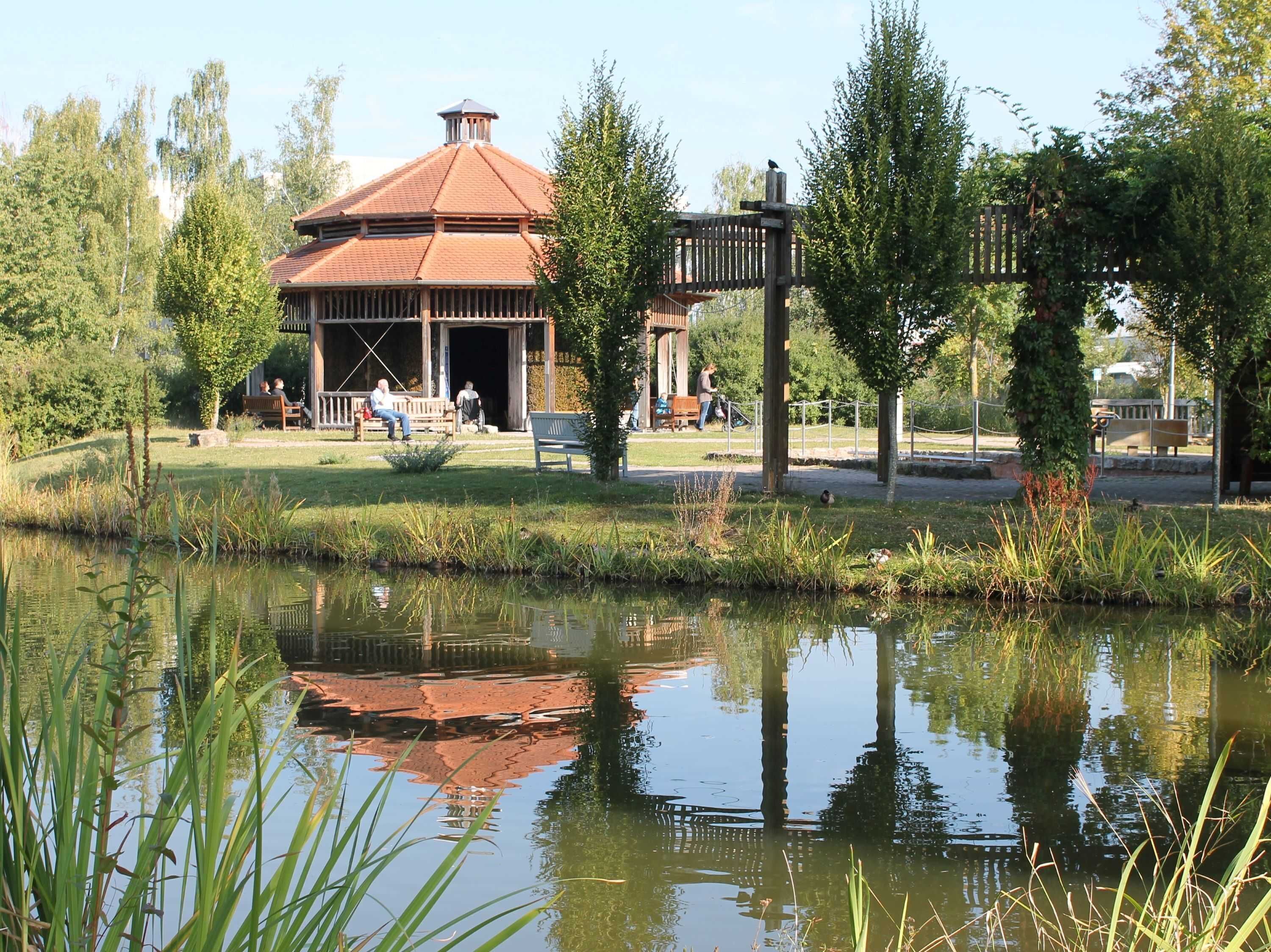Un pavillon doté d’un toit en tuiles, entouré d’une pelouse agrémentée de bancs et d’un étang dans lequel se reflète la structure.