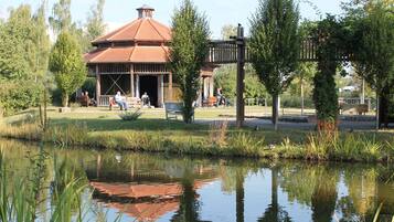 Un pavillon doté d’un toit en tuiles, entouré d’une pelouse agrémentée de bancs et d’un étang dans lequel se reflète la structure.