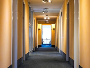 Un corridoio d'albergo con pavimento rivestito di moquette, soffitto con illuminazione a incasso e una finestra che offre vista sull'esterno.