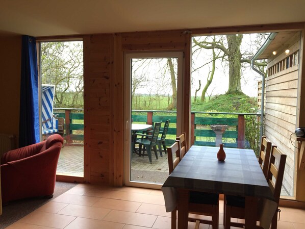 Un intérieur en bois avec une table et des chaises, un fauteuil rouge et une vue sur une terrasse verdoyante et des arbres à l’extérieur.