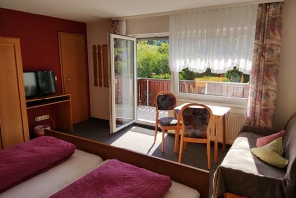 Une pièce comprenant un lit, un canapé, une télévision, une petite table et une chaise. Une porte-fenêtre coulissante donne accès à un balcon d’où l’on peut admirer la verdure.