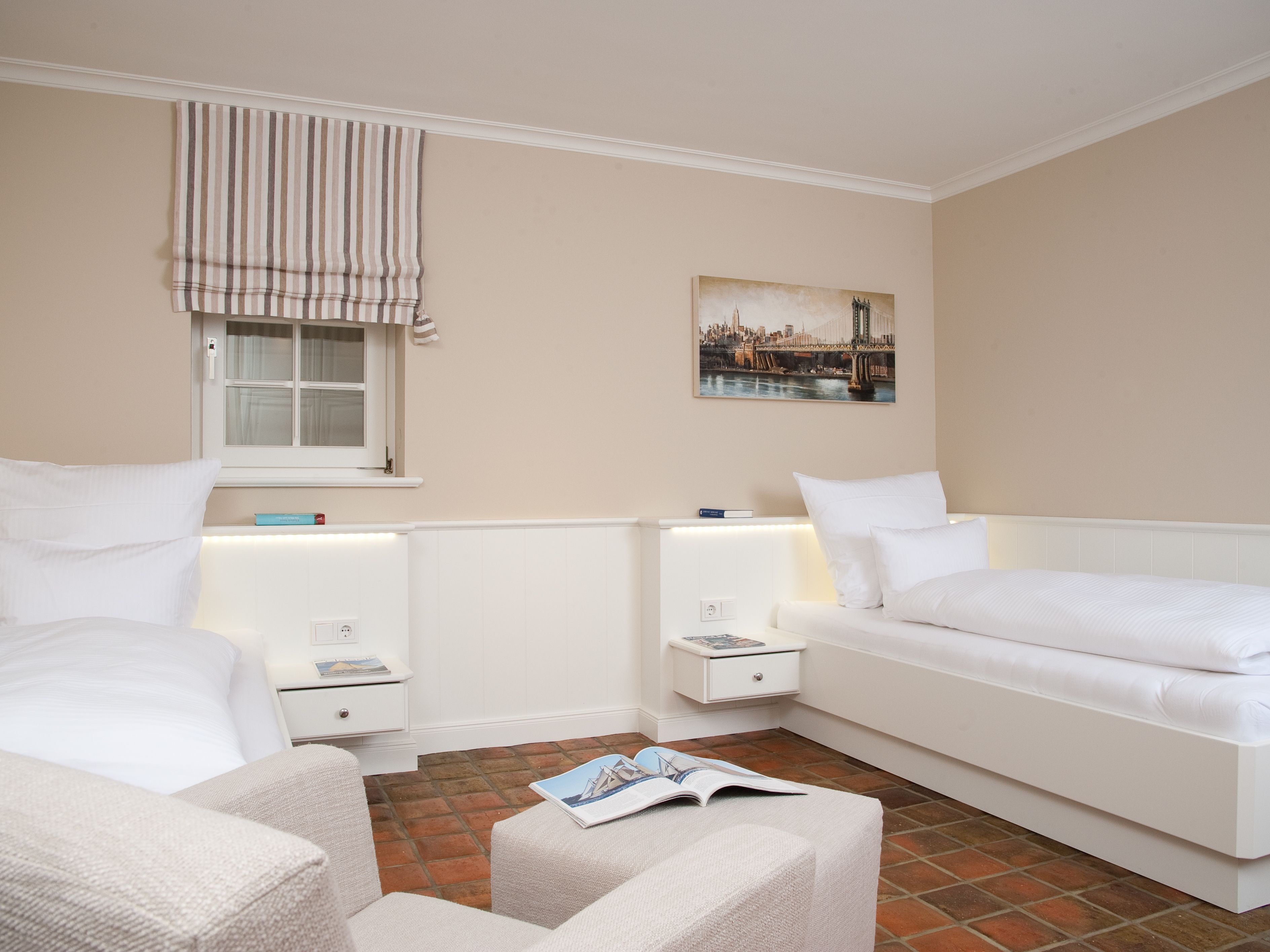 Habitació d'hotel amb dos llits individuals, una finestra amb cortines a ratlles, un quadre de paisatge urbà i un paviment de rajoles.