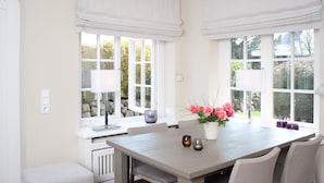 A dining area with a table, chairs, and a vase of flowers. There are windows with curtains and a view of greenery outside. - Vis à Vis from A&M - Vis à Vis (Kampen (Sylt))