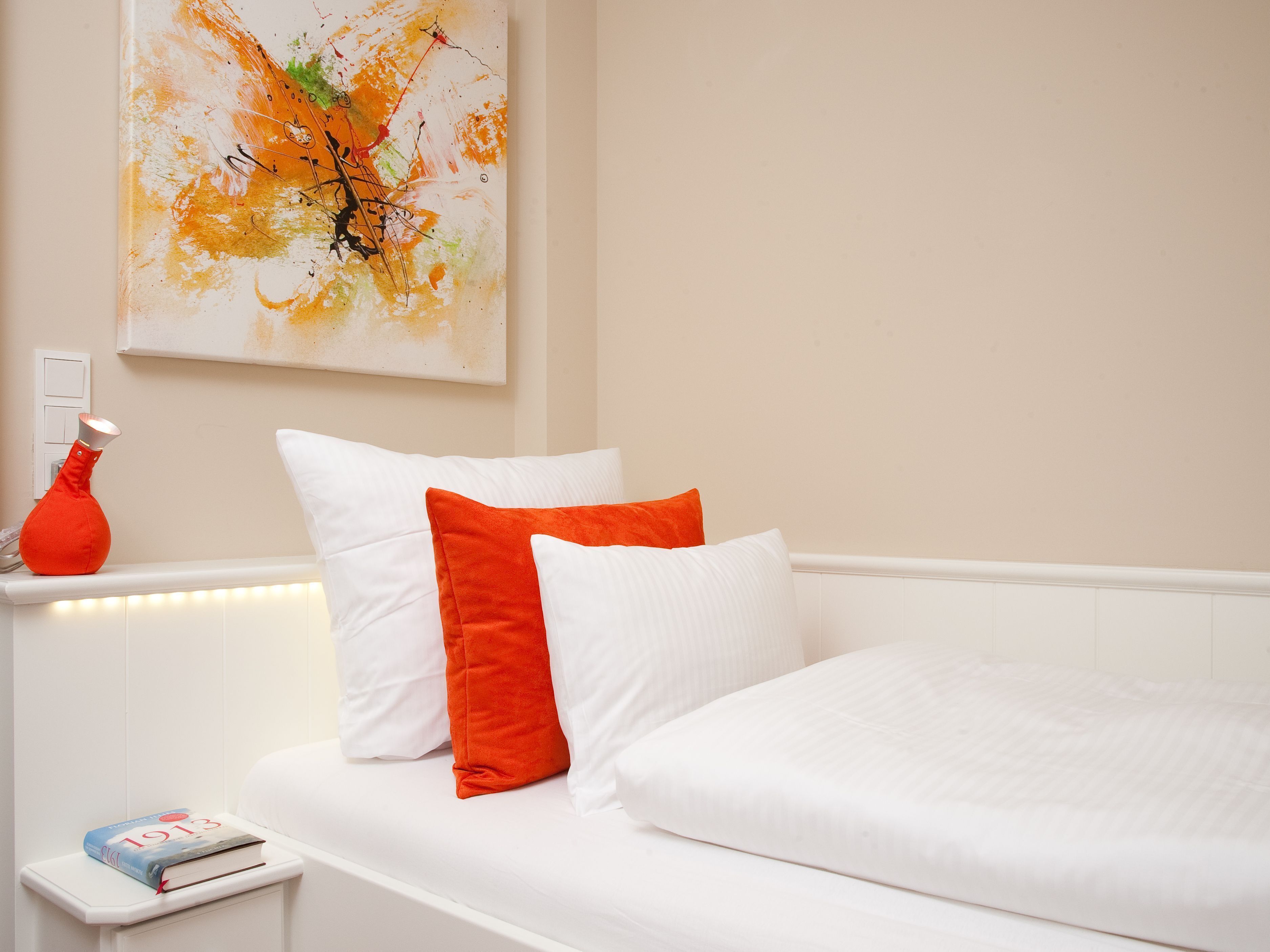 Una cama bien hecha con almohadas blancas y naranjas, un florero rojo sobre una mesita de noche y un cuadro abstracto colgado en la pared.