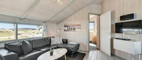 Een modern interieur met een zwarte leren bank, een klein rond tafeltje met een plant, en een keukenhoek met een televisie en apparatuur.