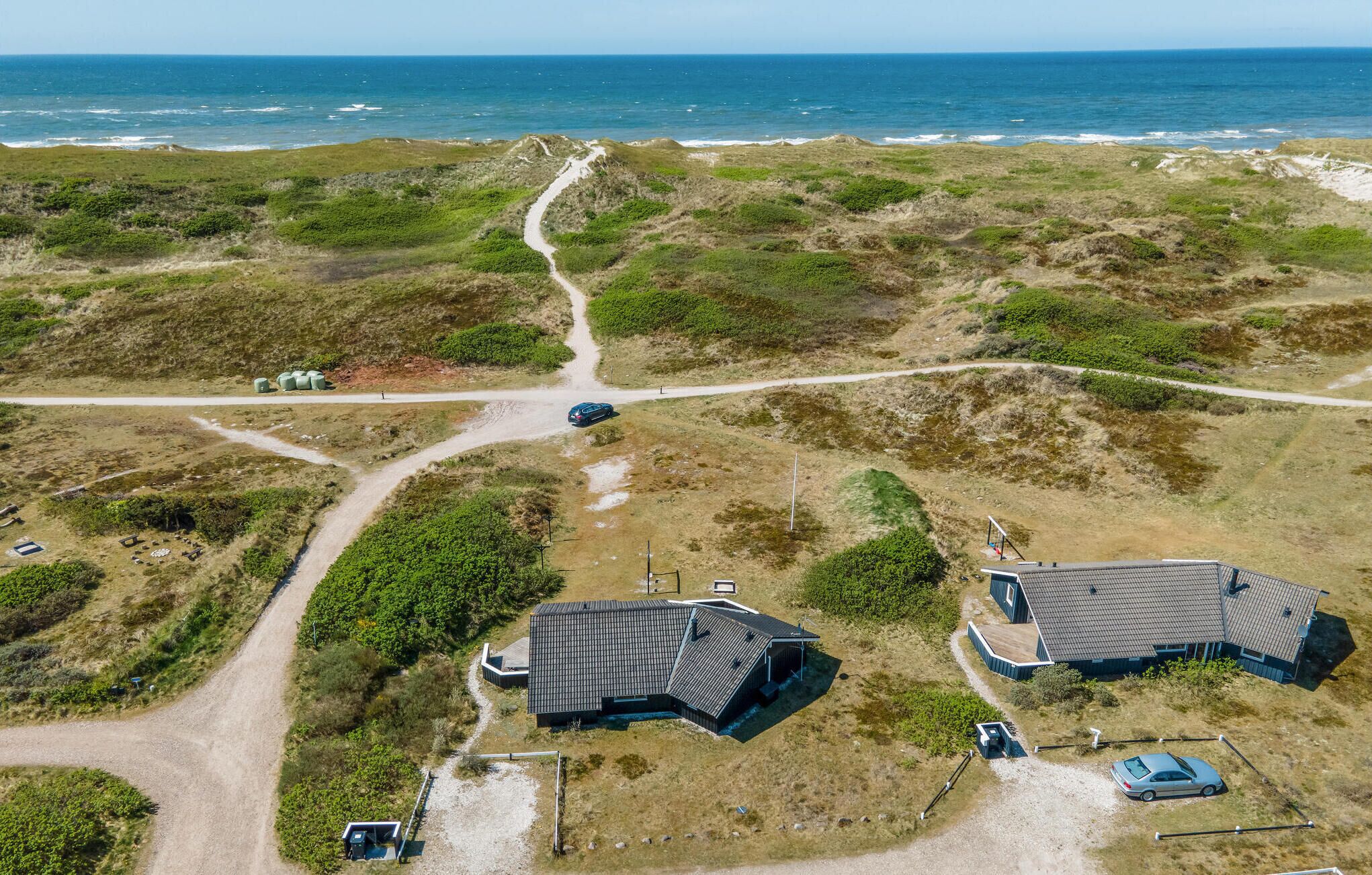 An aerial view of a coastal property with two main buildings, a dirt path, and a car parked near the structures.