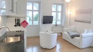 Un salon moderne avec un canapé blanc, un téléviseur à écran plat, une table à manger et un coin cuisine équipé d’un évier et de placards.