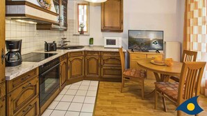 Een keuken met houten kastjes, een aanrecht, een kookplaat, een spoelbak, een magnetron, een televisie en een eettafel met stoelen.