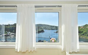 A room with a large window overlooking a body of water, a balcony with a railing, and a view of a hillside with buildings. - Cozy apartment in Straumsgrend (Straumsgrend)
