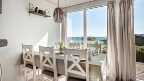 A dining area with a table and chairs, a hanging light fixture, and a view of the sea through large windows.