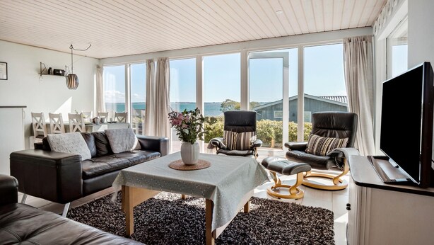 A spacious living room with a large window offering a view of the sea, a comfortable sofa, a dining area with chairs, and a television.