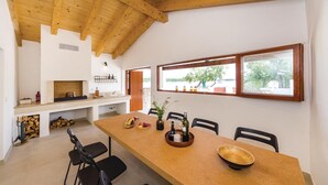 A modern kitchen with a wooden ceiling, a dining area with a long table, chairs, and a view of the outdoors through large windows.