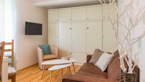 Una sala de estar con un sofá marrón, una mesa de centro blanca, un sillón beige y un televisor montado en la pared.