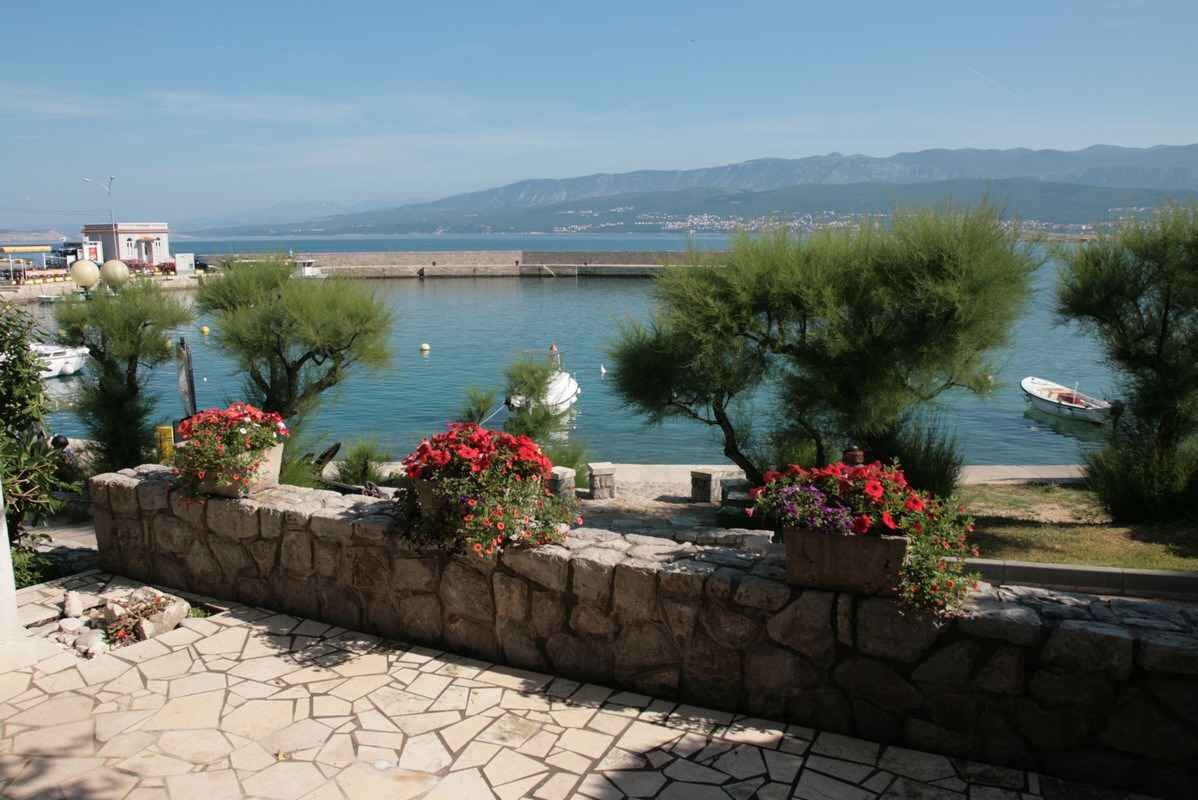 A coastal view with a stone wall, potted plants, and a clear blue sea.