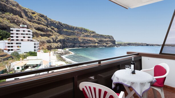 A balcony with a table and chairs overlooking a coastal view, including a building and rocky cliffs.