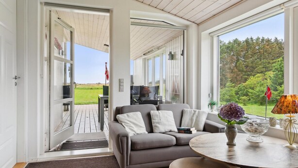 Sala moderna com sofá cinzento, mesa redonda e vista para um jardim através de grandes janelas.