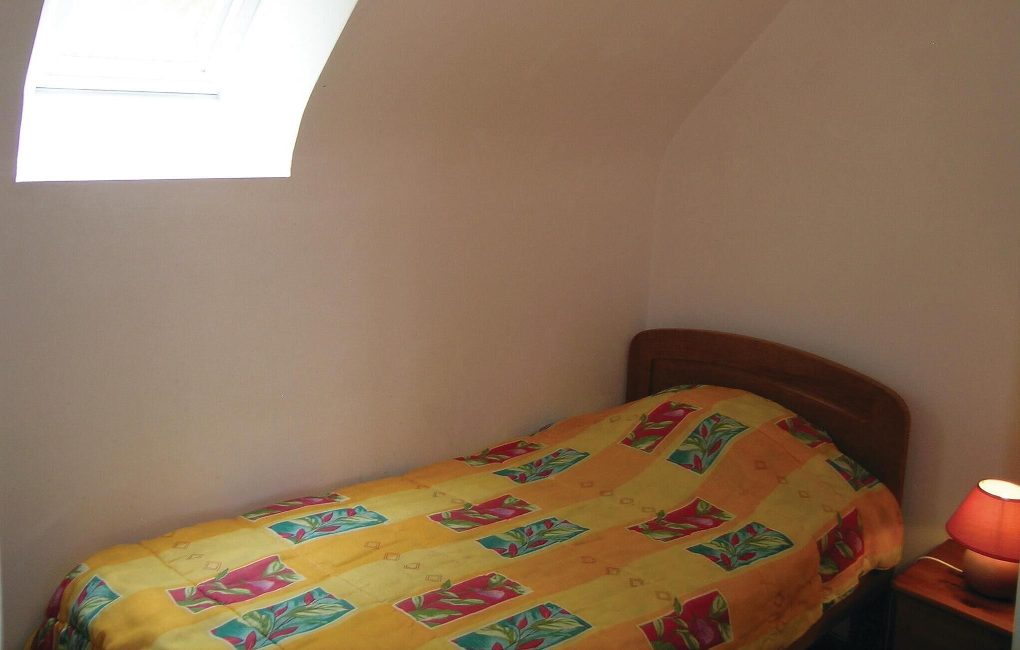 A small, single-bed room with a colorful bedspread, a nightstand with a lamp, and a window providing natural light.
