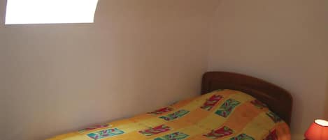 A small, single-bed room with a colorful bedspread, a nightstand with a lamp, and a window providing natural light.