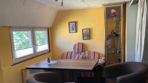 Ett mysigt rum med en säng, ett bord och två stolar. Det finns två fönster med persienner, ett element och en hylla med glasvaror.