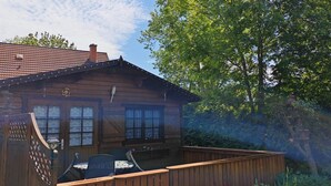 En stuga av trä med en veranda, omgiven av frodig grönska och blommande växter.