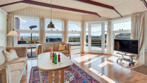 A spacious living area with a high ceiling, large windows offering a view of the sea, a sofa, a coffee table with bottles, and a flat-screen TV. - Stunning home in Sjølund with kitchen (Sjølund)