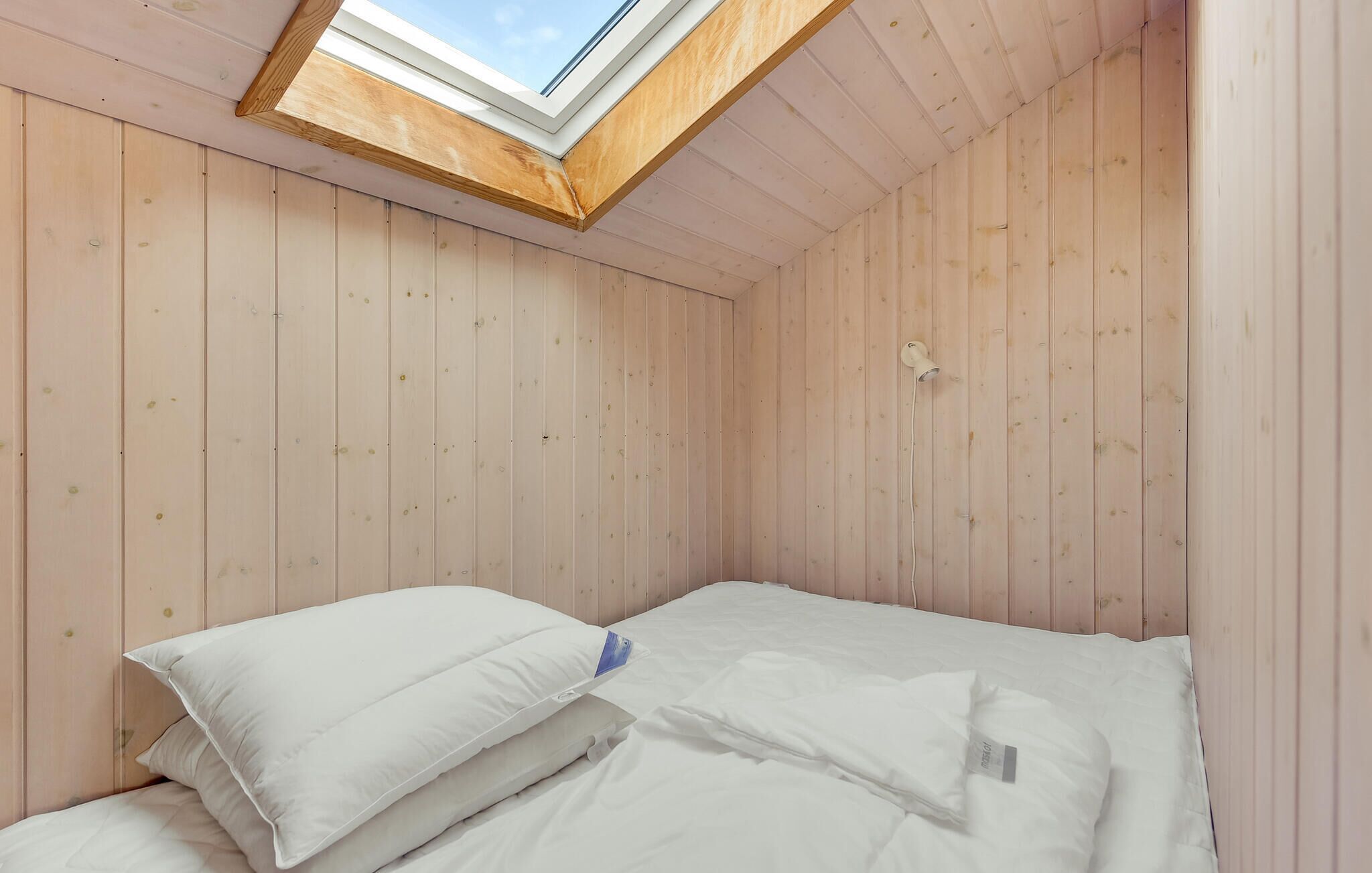 A small, cozy room with wooden walls, a skylight, and a neatly made bed with white bedding.