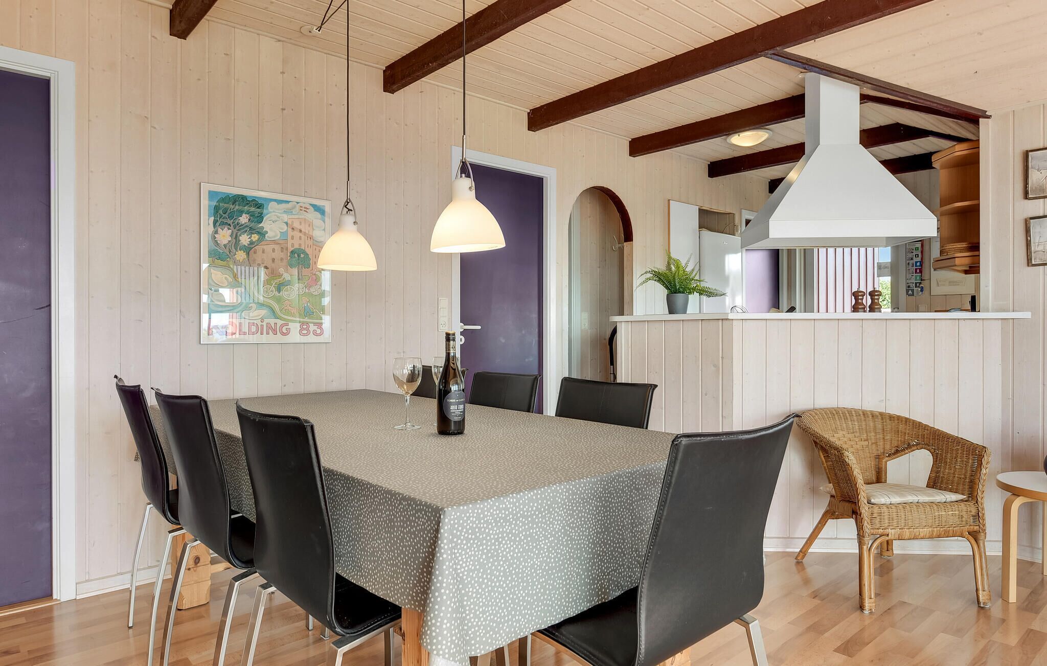 Un comedor con una mesa grande, sillas negras y una silla de mimbre. Hay lámparas colgantes sobre la mesa y una zona de cocina al fondo.