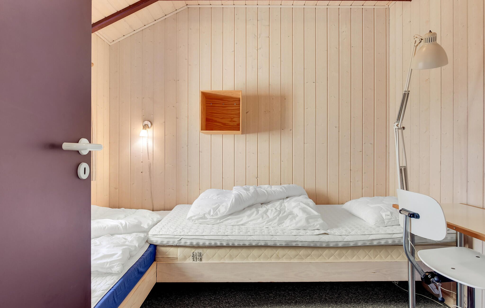 A room with a wooden bunk bed, a small wooden cabinet, a bedside table with a lamp, and a door with a handle.