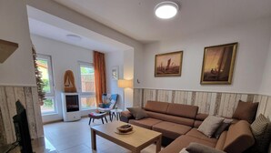 A living room with a sofa, a coffee table, a TV, and a fireplace. There are two paintings on the wall and a window with curtains.