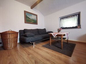 Un salon chaleureux avec un canapé gris foncé, une table basse en bois recouverte d’une nappe en dentelle, un panier en osier et un tableau représentant un paysage de montagne accroché au mur.