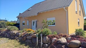 Une maison jaune avec un toit en tuiles, une porte et une fenêtre. On peut voir des rochers et des plantes devant la maison.