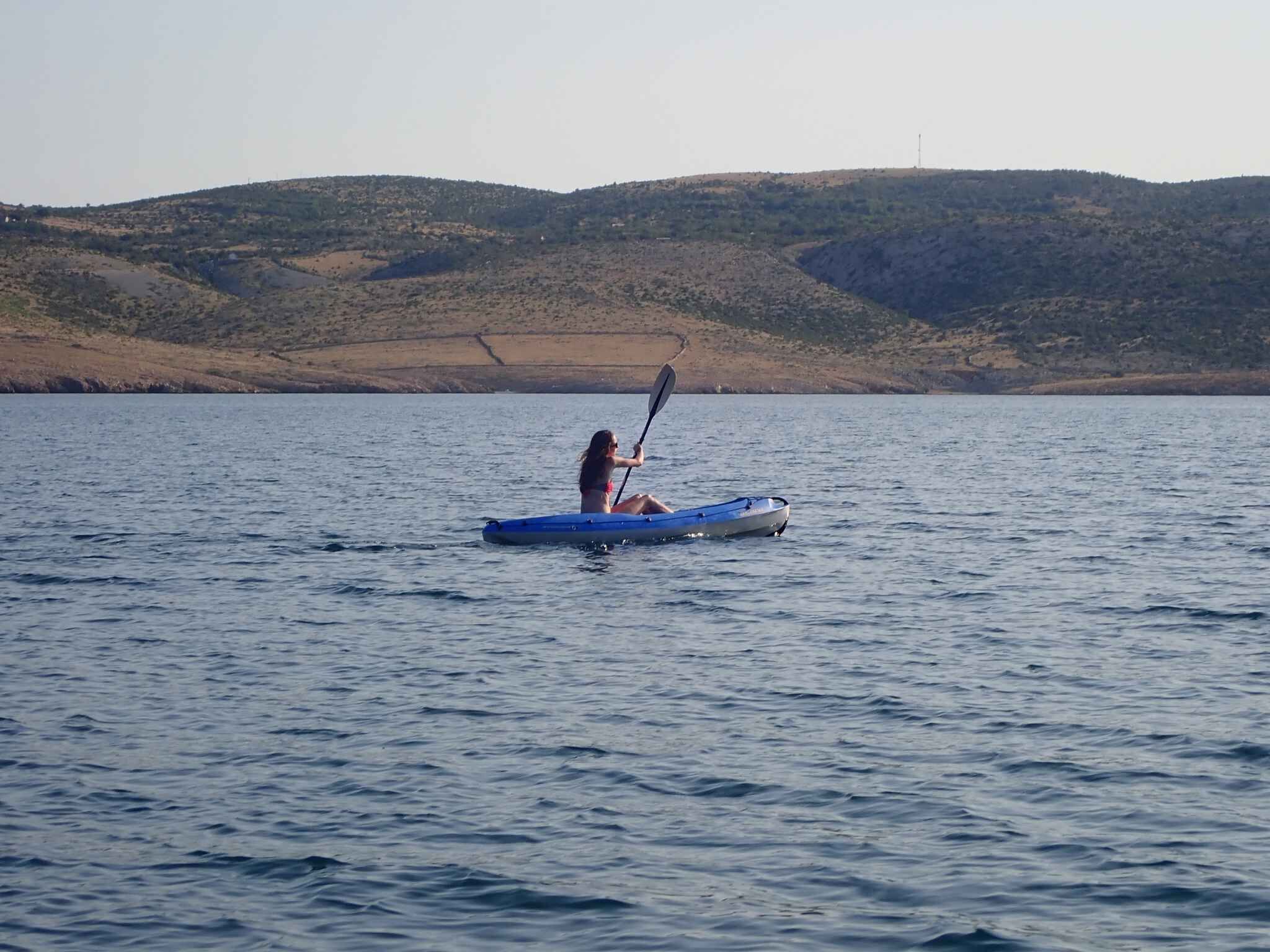 A person kayaking on a calm body of water with hills in the background.