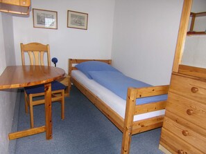 Acomodação pequena, em um único quarto, com uma cama de madeira, uma escrivaninha de madeira e uma cadeira.