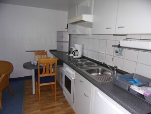 A compact kitchen with a stove, sink, and a coffee maker. There is a dining table with chairs and a wicker chair.